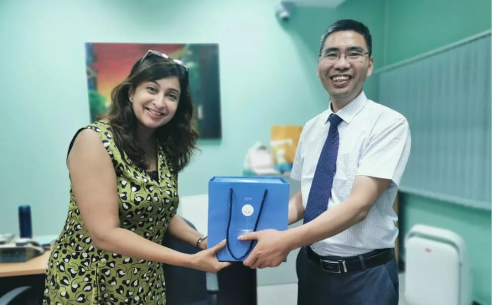 Two smiling people hold a blue gift bag together in a teal office room.